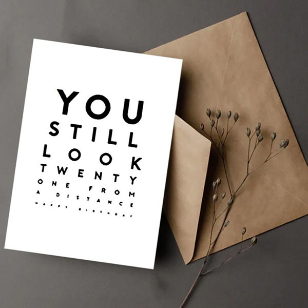 Greeting card with text 'YOU STILL LOOK TWENTY ONE FROM A DISTANCE' next to a brown envelope on a gray background.